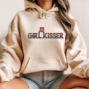 Puede incluir: Sudadera beige con bolsillo delantero y cordón. La sudadera presenta el texto "GIRL KISSER" en colores arcoíris. Encima del texto hay un gráfico de dos figuras besándose, también en colores arcoíris.