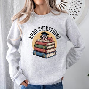 Op de afbeelding: Een lichtgrijs sweatshirt met de tekst "READ EVERYTHING". De afbeelding toont een cartoonworm met een bril en een afstudeerhoed, zittend op een stapel kleurrijke boeken. De boeken zijn rood, groen en blauw.