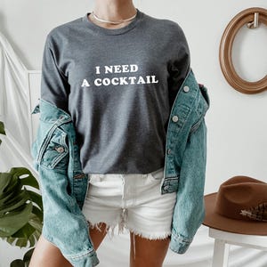 May include: A charcoal gray long-sleeve shirt with the white text "I NEED A COCKTAIL". The shirt is paired with white shorts and a light blue denim jacket. A brown hat sits on a white stool in the background.