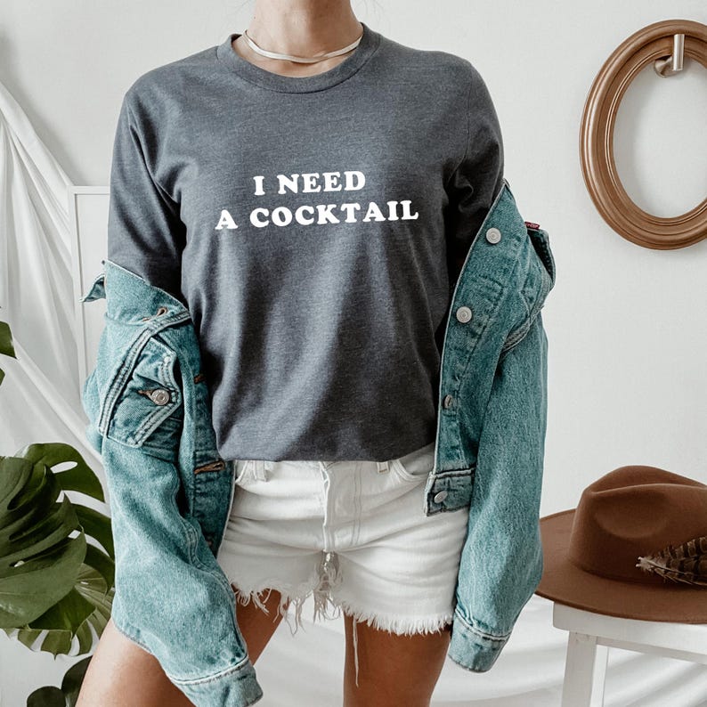 May include: A charcoal gray long-sleeve shirt with the white text "I NEED A COCKTAIL". The shirt is paired with white shorts and a light blue denim jacket. A brown hat sits on a white stool in the background.