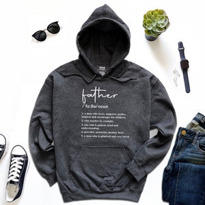 K&ouml;nnte beinhalten: Ein dunkelgrauer Kapuzenpullover mit der Definition von "Vater" in Wei&szlig; aufgedruckt. Die Definition enth&auml;lt die phonetische Aussprache und f&uuml;nf beschreibende Punkte. Der Pullover wird mit Sonnenbrille, Uhr und Jeans pr&auml;sentiert.