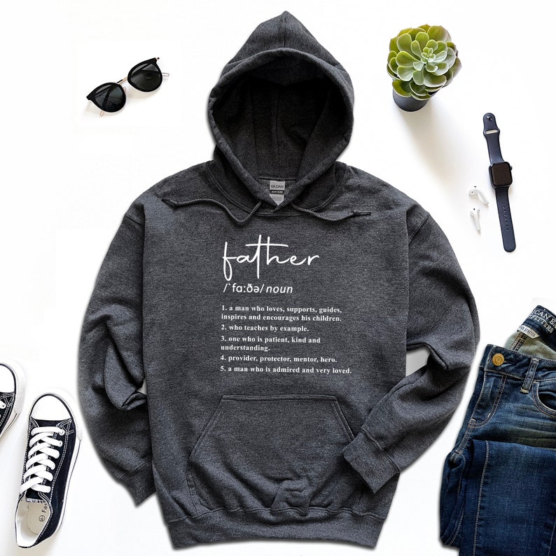 K&ouml;nnte beinhalten: Ein dunkelgrauer Kapuzenpullover mit der Definition von "Vater" in Wei&szlig; aufgedruckt. Die Definition enth&auml;lt die phonetische Aussprache und f&uuml;nf beschreibende Punkte. Der Pullover wird mit Sonnenbrille, Uhr und Jeans pr&auml;sentiert.