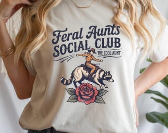 May include: Cream-colored t-shirt with a graphic featuring the text "Feral Aunts SOCIAL CLUB" and "THE COOL AUNT." The graphic depicts a person riding a raccoon, with a rose at the bottom. The design is in shades of blue, brown, and red.