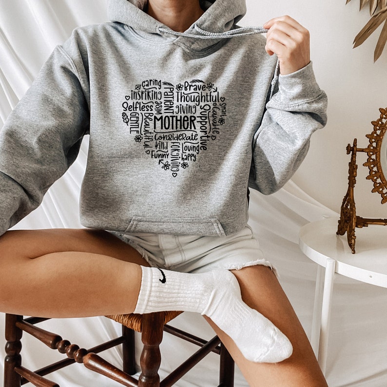 Op de afbeelding: Een grijs hoodie met een zwart hartvormig ontwerp met het woord "MOEDER" omringd door positieve adjectieven.