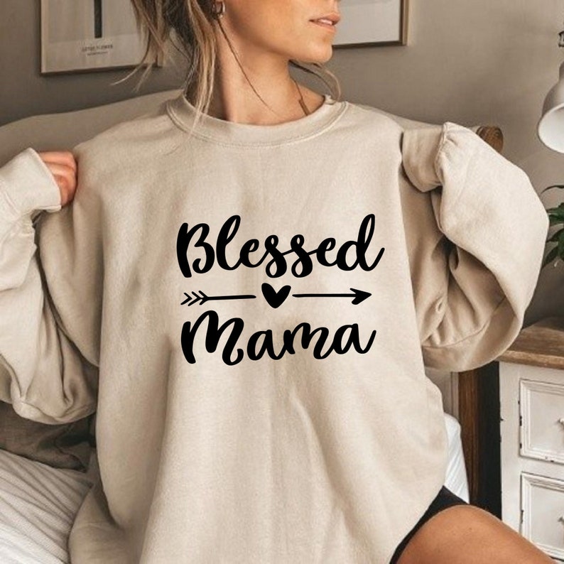 Puede incluir: Una sudadera beige con un gr&aacute;fico negro que dice "Blessed Mama" con un dise&ntilde;o de flecha y coraz&oacute;n.
