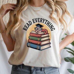 Może przedstawiać: Kremowa koszulka z grafiką mola książkowego w okularach i czapce absolwenta, siedzącego na stosie książek. Tekst "READ EVERYTHING" jest wygięty nad ilustracją, na żółtym okręgu.