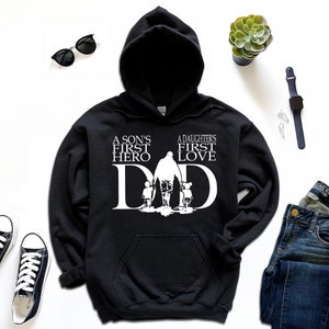 May include: Black hooded sweatshirt with a white graphic design. The graphic features a father walking with a son and daughter. The text reads "A son's first hero, A daughter's first love, DAD".