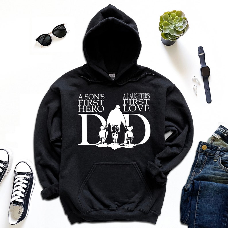 May include: Black hooded sweatshirt with a white graphic design. The graphic features a father walking with a son and daughter. The text reads "A son's first hero, A daughter's first love, DAD".