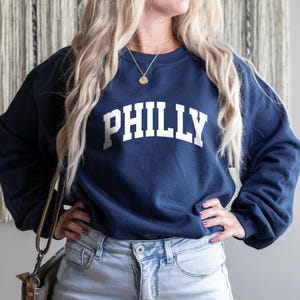 Könnte beinhalten: Ein marineblauer Pullover mit dem Wort "PHILLY" in weiß auf der Vorderseite. Der Pullover wird von einer Person mit langen blonden Haaren getragen und zu einer hellblauen Jeans kombiniert.