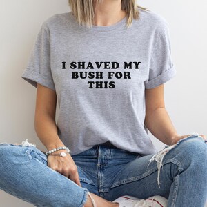 May include: A heather gray t-shirt with the text "I shaved my bush for this" in black block letters. The shirt has short sleeves and a relaxed fit. The model is wearing blue jeans and white sneakers.