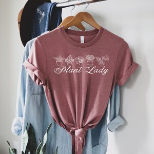 K&ouml;nnte beinhalten: Ein mauvefarbenes T-Shirt mit einer wei&szlig;en Grafik von Topfpflanzen und dem Text "Plant Lady".