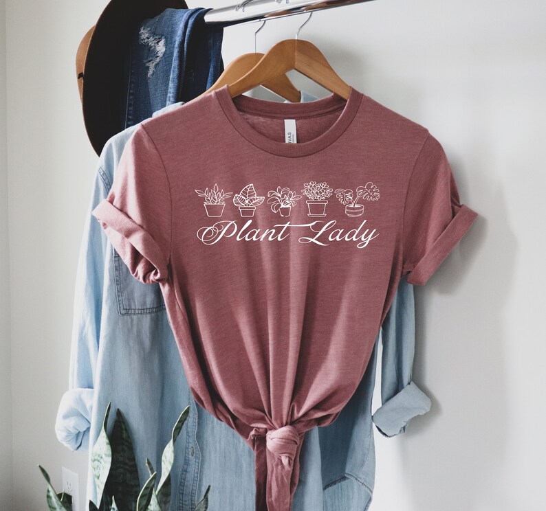 K&ouml;nnte beinhalten: Ein mauvefarbenes T-Shirt mit einer wei&szlig;en Grafik von Topfpflanzen und dem Text "Plant Lady".