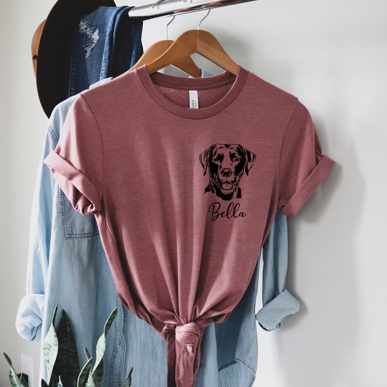 May include: A maroon t-shirt with a black silhouette of a Labrador Retriever and the name "Bella" printed on the front.