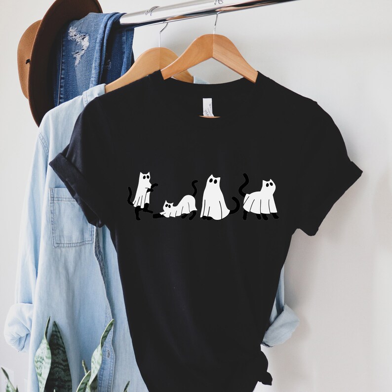 May include: Black t-shirt with a graphic of five white cats dressed as ghosts. The cats are in a line, with their tails trailing behind them.