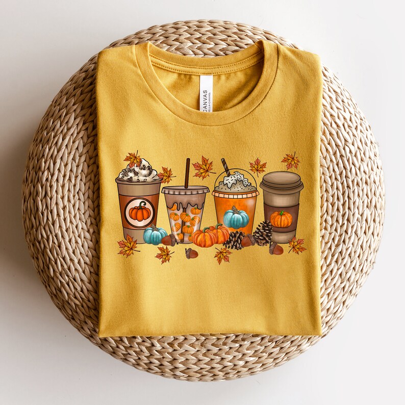 May include: A mustard yellow t-shirt featuring a graphic of four autumn-themed coffee drinks. Each drink is decorated with a pumpkin, autumn leaves, and pine cones.