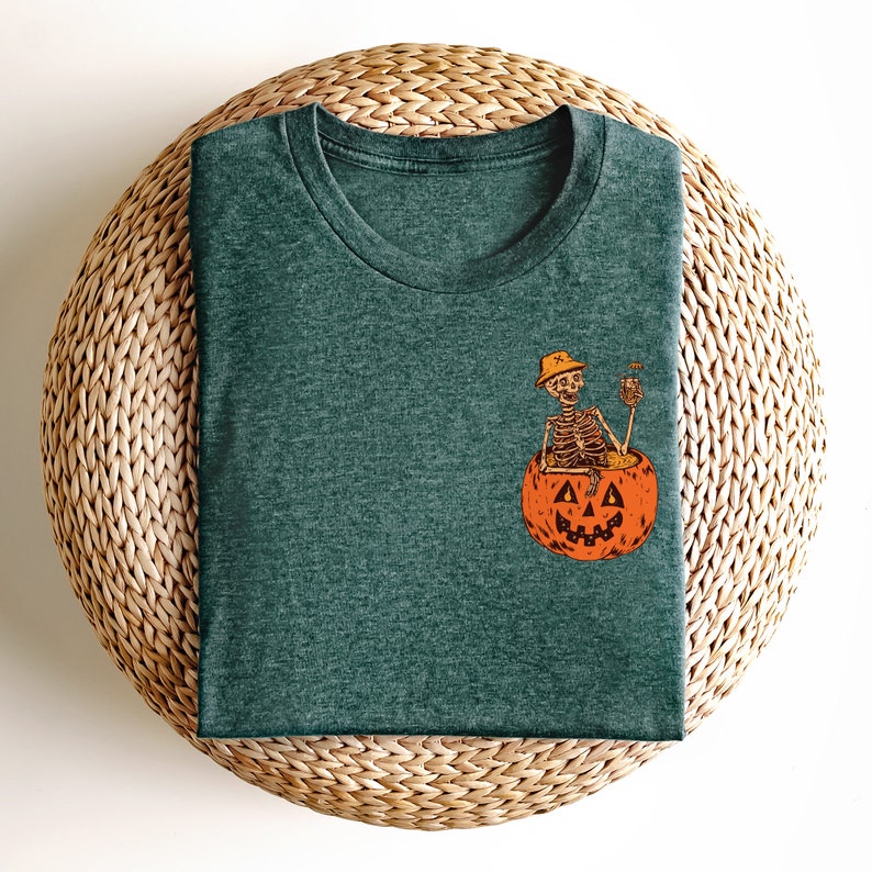 May include: A dark green t-shirt with a graphic of a skeleton sitting inside a jack-o'-lantern holding a drink. The skeleton is wearing a hat.