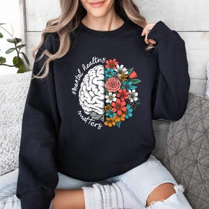 Może przedstawiać: Granatowa bluza z napisem "Mental Health Matters" oraz grafiką mózgu i kwiatów. Mózg jest biały, a kwiaty czerwone, pomarańczowe, niebieskie i białe.