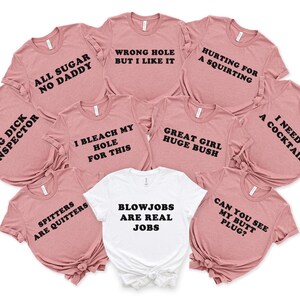 May include: A collection of pink t-shirts with various humorous phrases printed in black, including "All Sugar No Daddy" and "I Need a Cocktail." One white t-shirt features the text "Blowjobs are Real Jobs."