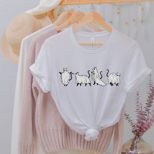May include: White t-shirt with a graphic of four cats dressed as ghosts. The cats are in a line, facing forward, and each has a different pose. The graphic is in black and white.