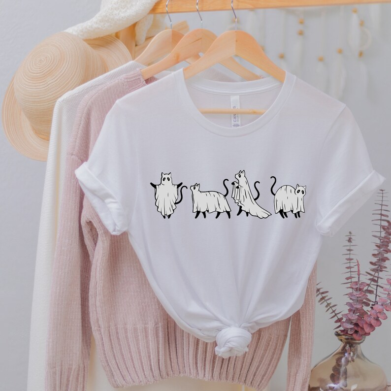 May include: White t-shirt with a graphic of four cats dressed as ghosts. The cats are in a line, facing forward, and each has a different pose. The graphic is in black and white.