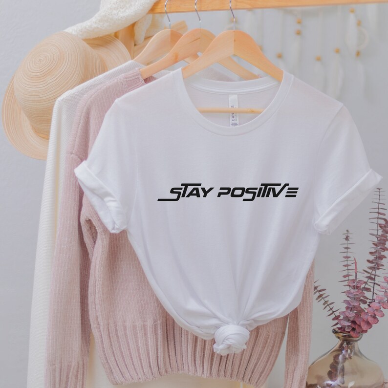 Puede incluir: Camiseta blanca con texto negro que dice "STAY POSITIVE" en una fuente estilizada.
