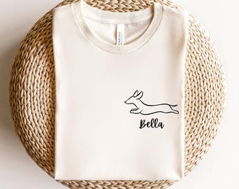 May include: Cream-colored t-shirt featuring a black line drawing of a dachshund dog in motion. The dog's name, "Bella," is written in a cursive font below the illustration. The shirt is folded and placed on a woven, circular surface.