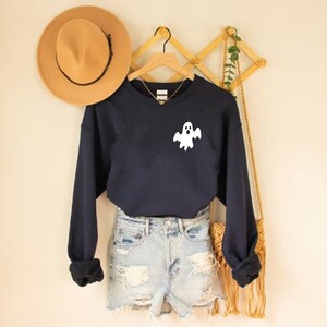 May include: A navy blue sweatshirt with a white ghost graphic on the front. The sweatshirt is hanging on a wooden hanger and is paired with denim shorts.