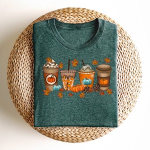 May include: A teal green t-shirt featuring a graphic of four autumn-themed coffee drinks with whipped cream, pumpkin spice, and autumn leaves. The drinks are surrounded by pumpkins, pine cones, and acorns.