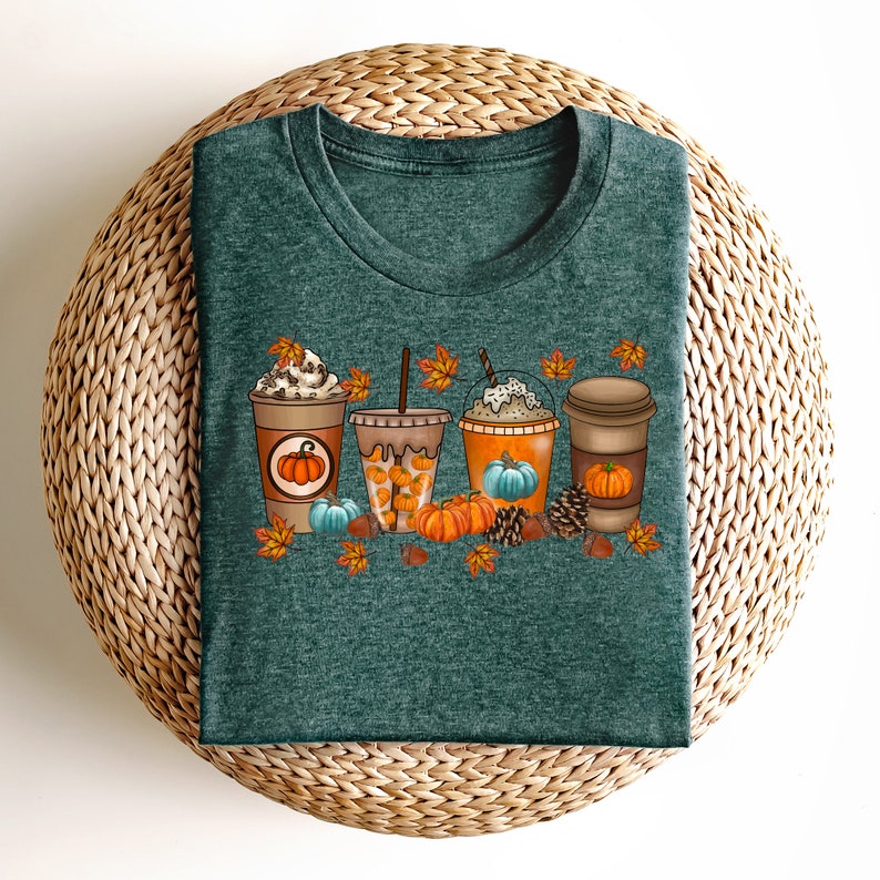 May include: A teal green t-shirt featuring a graphic of four autumn-themed coffee drinks with whipped cream, pumpkin spice, and autumn leaves. The drinks are surrounded by pumpkins, pine cones, and acorns.
