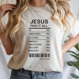 Puede incluir: Una camiseta beige con el texto negro "JESUS PAID IT ALL." Debajo, una lista de pecados como "SIN" y "ANGER & HATE" se emparejan con "PAID." Un código de barras y "Romanos 6:23" están en la parte inferior.