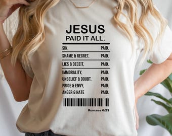 Könnte beinhalten: Ein beiges T-Shirt mit dem schwarzen Text "JESUS PAID IT ALL." Darunter eine Liste von Sünden wie "SIN" und "ANGER & HATE", die mit "PAID" gepaart sind. Ein Barcode und "Römer 6:23" befinden sich unten.