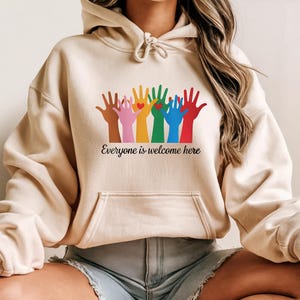Op de afbeelding: Beige hoodie met een voorzak en trekkoord. De voorkant heeft een afbeelding van kleurrijke handen met een hart en de tekst "Everyone is welcome here" in het zwart.