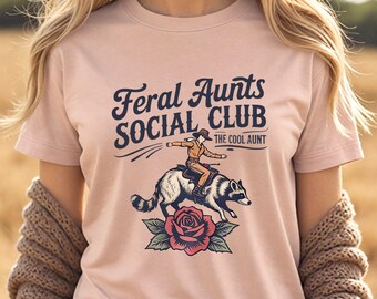 May include: A light pink t-shirt with the text "Feral Aunts SOCIAL CLUB" and an illustration of a person riding a raccoon over a rose. The text "THE COOL AUNT" is below the main text.