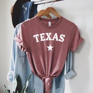 May include: A mauve colored t-shirt with the word "TEXAS" in white with a white star underneath. The shirt is tied at the bottom.