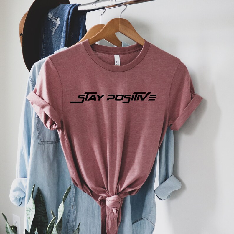 Puede incluir: Una camiseta de color malva con mangas cortas y cuello redondo. La camiseta tiene el texto "STAY POSITIVE" impreso en negro en la parte delantera.