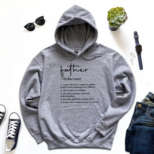 K&ouml;nnte beinhalten: Grauer Kapuzenpullover mit dem Wort "father" und seiner Definition in schwarzer Schrift. Die Definition beschreibt einen liebevollen, unterst&uuml;tzenden Mann. Das Bild zeigt auch Jeans, Sonnenbrille und Turnschuhe, f&uuml;r einen l&auml;ssigen Look.