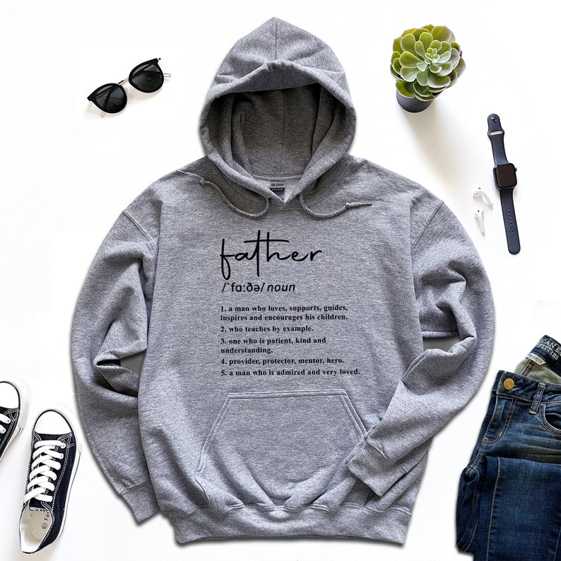 K&ouml;nnte beinhalten: Grauer Kapuzenpullover mit dem Wort "father" und seiner Definition in schwarzer Schrift. Die Definition beschreibt einen liebevollen, unterst&uuml;tzenden Mann. Das Bild zeigt auch Jeans, Sonnenbrille und Turnschuhe, f&uuml;r einen l&auml;ssigen Look.