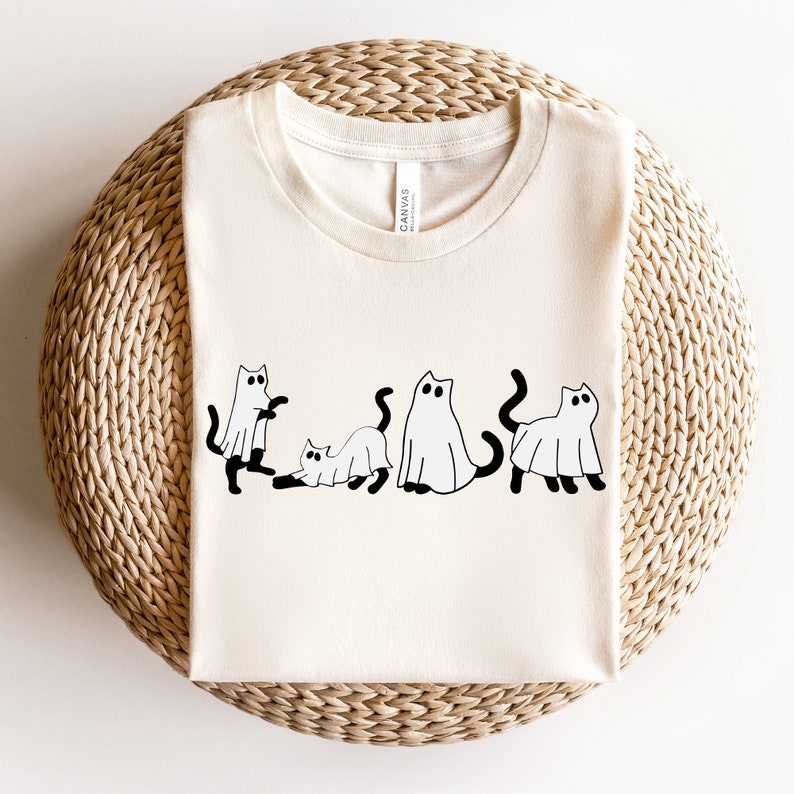 May include: A white t-shirt with a black graphic of five cats dressed as ghosts. The cats are in a line, with the first cat looking to the left, the second cat looking to the right, the third cat looking to the left, the fourth cat looking to the right, and the fifth cat looking to the left. The graphic is simple and minimalist, with the cats drawn in a cartoon style.