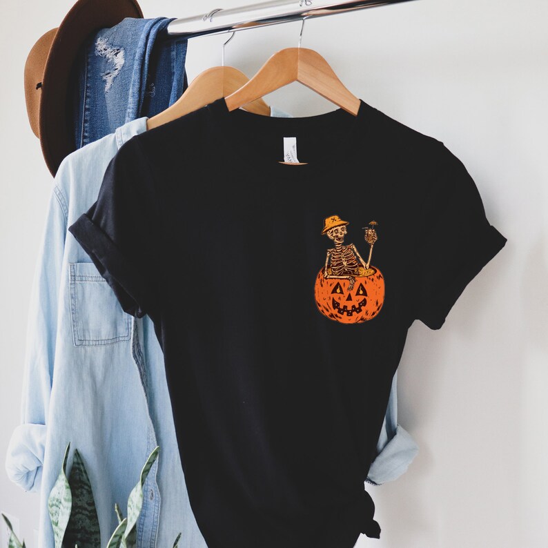 May include: Black t-shirt with a graphic of a skeleton sitting in a jack-o'-lantern holding a drink. The skeleton is wearing a hat.