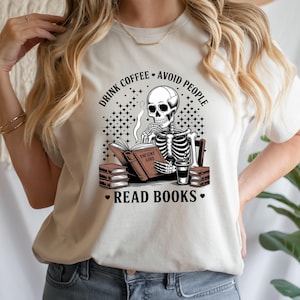 Op de afbeelding: Lichtgekleurd T-shirt met een skelet dat een boek leest met de tekst "Ancient Lore". Het shirt heeft ook de zinnen "Drink Coffee, Avoid People, Read Books". Een koffiekopje en stapels boeken zijn ook zichtbaar.