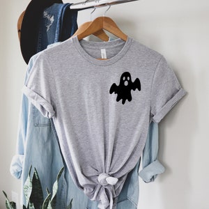 以下が含まれることがあります： 前面に黒い幽霊のグラフィックがプリントされたグレーのTシャツ。幽霊は驚いた表情をして、白いシーツを着ています。