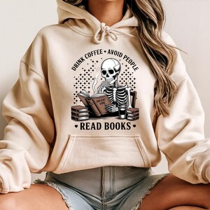Op de afbeelding: Beige hoodie met een voorzak en trekkoord. De afbeelding toont een skelet dat een boek leest met de tekst "DRINK COFFEE • AVOID PEOPLE • READ BOOKS." Het skelet rookt en heeft een koffiekopje ernaast. Het boek heet "ANCIENT LORE."