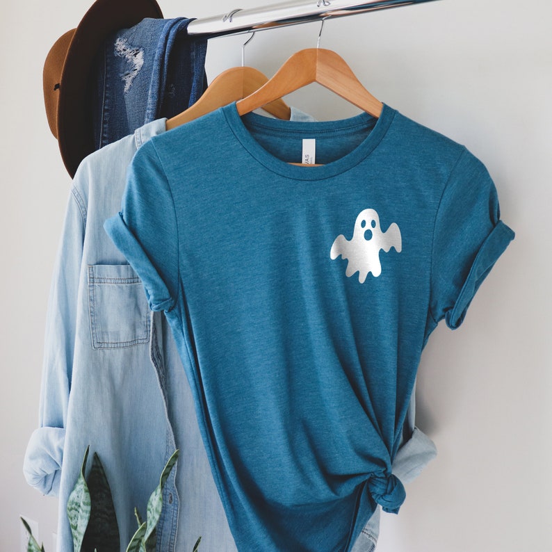以下が含まれることがあります： 前面に白い幽霊のグラフィックが描かれたターコイズブルーのTシャツ。Tシャツは木製ハンガーにかけられています。
