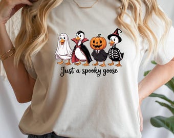 May include: A cream-coloured t-shirt with a Halloween design. The graphic depicts four cartoon geese in costumes: a ghost, a vampire, a pumpkin, and a skeleton. The text "Just a spooky goose" is printed below the geese.