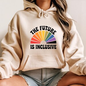 May include: A beige hoodie with a front pocket and drawstring. The front features a rainbow-colored design in a semi-circle with the words "THE FUTURE IS INCLUSIVE" in black, arched above and below the rainbow.
