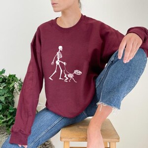 Pode incluir: Uma camisa de frio vinho com um gr&aacute;fico branco de um esqueleto passeando um esqueleto de cachorro.