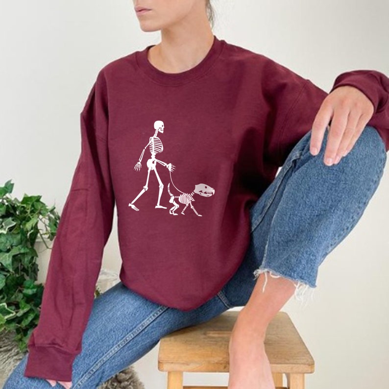 Pode incluir: Uma camisa de frio vinho com um gr&aacute;fico branco de um esqueleto passeando um esqueleto de cachorro.