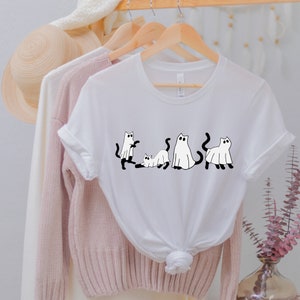 May include: White t-shirt with a black and white graphic of four cats dressed as ghosts. The cats are in different poses, including standing, sitting, and lying down.