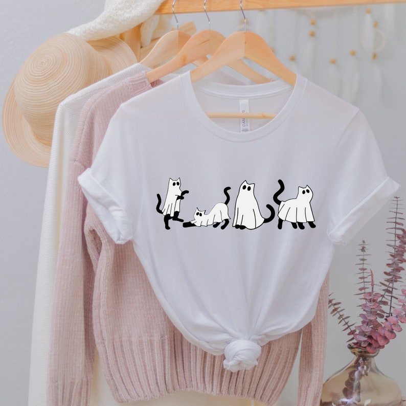 May include: White t-shirt with a black and white graphic of four cats dressed as ghosts. The cats are in different poses, including standing, sitting, and lying down.