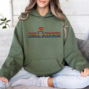May include: An olive green hoodie with a front pocket and a hood. The hoodie features the text "GIRL KISSER" in rainbow colors. The design includes a rainbow-colored graphic above the text.